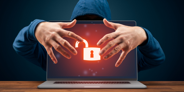 hooded man behind computer with face hidden, has hands in front of an unlocked computer screen as if he is hacking into it